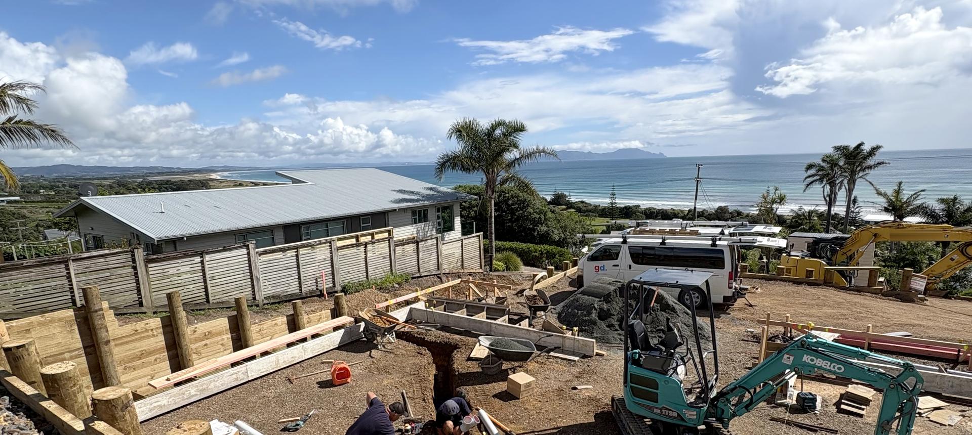 A GD Plumbing Services crew laying under slab drainage in the foundation trenches of a new build above the coast, with a branded van and a digger on site and the sea behind.